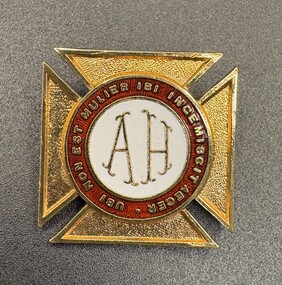 A gold and white enamel badge with the letters "AH" in the centre, surrounded by a red ring with Latin text, on a textured white background.