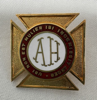 A gold and white enamel badge with the letters "AH" in the centre, surrounded by a red ring with Latin text, on a textured white background.