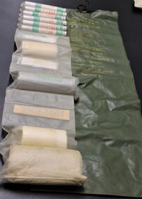 A long khaki outer sleeve that can be rolled and tied has list of contents and instructions for use in yellow. Inner sleeve is sectioned and contains tubes of pills, ointments, and bandages.