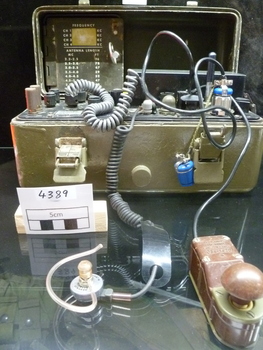 A radio set contained in an olive drab metal case, containing an ear piece on spiral cord and a bakelite morse code key, a lid listing antenna length and frequency channels. The base has switches, dials and a battery.