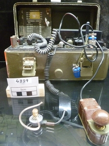 A radio set contained in an olive drab metal case, containing an ear piece on spiral cord and a bakelite morse code key, a lid listing antenna length and frequency channels. The base has switches, dials and a battery.