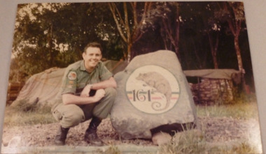 A colour photograph of WO2 Robert Palmer Matthews squatting beside 'Possum Rock' at Nui Dat.