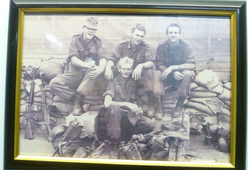 Black and white photograph depicting four soldiers seated on sandbag with their packs and guns