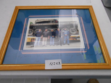 Coloured photograph backed on blue cardboard of five men in front of a tank which is loaded on a low loader.