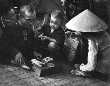 A photograph of a badly deformed male Vietnamese lottery ticket seller and his son with a very pronounced hair lip selling tickets to a female customer.