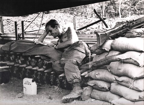 A photograph of a gunner of 104 Battery resting on a stack of 105mm shells reading a news sheet during a break in operations.