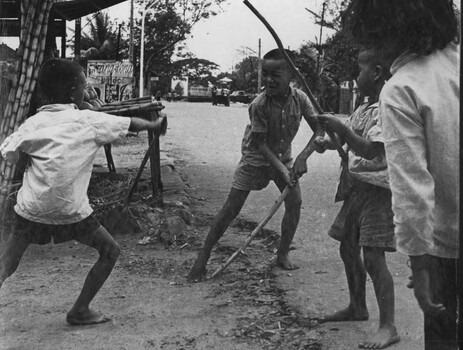 A photograph of In Baria, Phuoc Tuy Province two Vietnamese boys play a martial arts games using sticks. A younger brother and sister look on