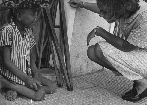 A photograph of an American Red Cross girl commonly called 'Doughnut Dollies' by American G.I's talks to a crippled Vietnamese street child.  