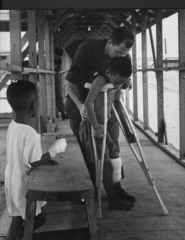 A photograph of an American Hospital Orderly assist a Vietnamese boy in how to use crutches after suffering a serious leg wound.