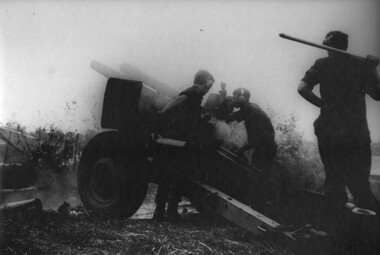 A Gunner at Fire Base Betty flicks a 105mm round casing behind him another adjusts the gunsight while a fellow gunner looks on. 