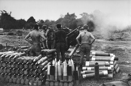 Six soldiers standing amongst artillary on the ground near a howitzer.