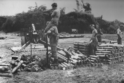 There are three soldiers standing amongst artllery shells. Also there is a soldier standing on round cylinders with another soldier on his shoulder.