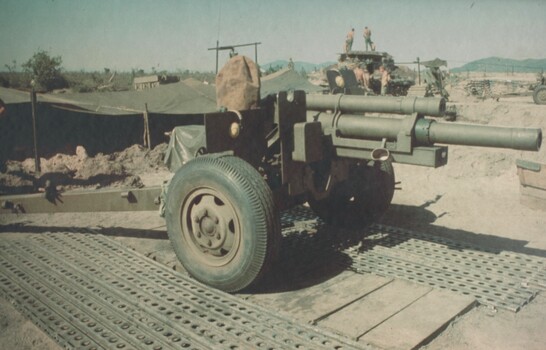 A photograph of a 105mm gun, sits in its bay at a Fire support Base at the base of the Nui Thai Vai Mountains. 