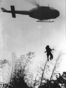 A photograph of an RAAF Huey 'slick' crew member steadying the winch wire as a wounded Digger from 8 RAR is medivaced. 