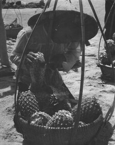 A photograph of a young Vietnamese girl prepares a pineapple for a serviceman on the sand at Back Beach, Vung Tau. 