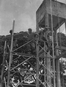 A black and white photograph of engineers constructing a Southern Cross Windmill tower next to the water header tower in Hoi My. 