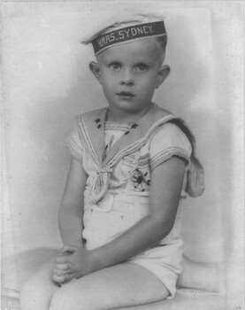 A black and white photograph of Denis Stanley Gibbons in 1940, aged three years proudly wearing a HMAS Sydney cap.