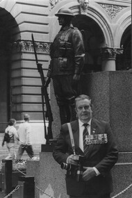 A photograph of Denis Gibbons, in 2004 standing at the Cenotaph in Martin Place, Sydney with his Nikon cameras and his Vietnam War decorations.