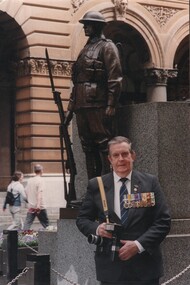 A colour photograph of Denis Gibbons, in 2004 standing at the Cenotaph in Martin Place, Sydney with his Nikon cameras and his Vietnam War decorations.