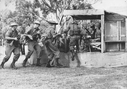 A photograph of assembled Infantry Diggers receiving instructions using a wood and hessian helicopter 'mockup' to practice embarking and disembarking. 