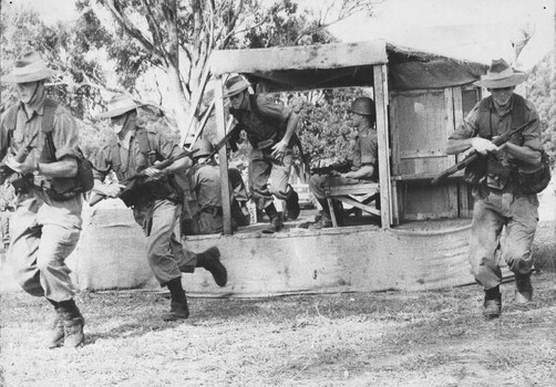 A photograph of assembled Infantry Diggers receiving instructions using a wood and hessian helicopter 'mockup' to practice embarking and disembarking. 