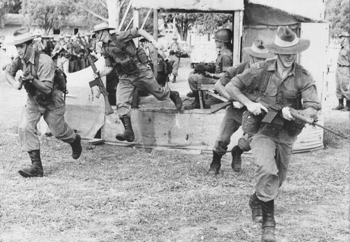 A photograph of assembled Infantry Diggers receiving instructions using a wood and hessian helicopter 'mockup' to practice embarking and disembarking. 