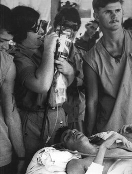 A photograph of Medics Rob Swincer and Mick O'Halloran steady a stretcher as Capt Ann Hall adjusts the drip bottle for a wounded Digger. 