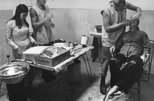 A photograph of a dentist works on a Vietnamese patient while a dental assistant and female Vietnamese aide look on. 