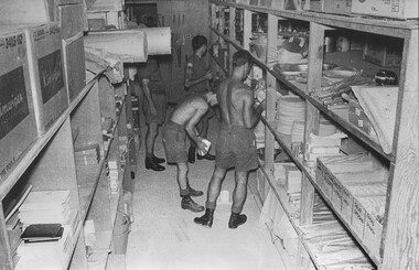 A photograph at the Main Quartermasters Store at 1st Australian Field Hospital, two storemen fill a requisition Capt John Knowles (partly obscured) watches.