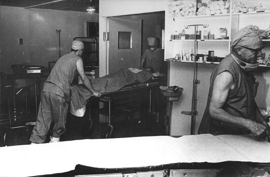 A photograph at 1st Australian Field Hospital, Theatre Sister June Minchow assisted by a theatre Medic wheels an American pilot to the ward. 