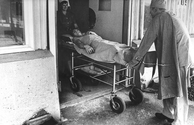 A photograph at the 1st Australian Field Hospital, Sister Lt Desley McLean assisted by a Medic wheels a patient out of intensive care ward. 