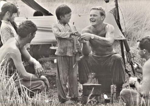 A photograph taken at Phuoc Tuy Province. Vietnamese children help 2Lt Mike Deak & Cpl Bob Kearney of 5 RAR prepare their evening meal.