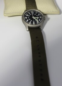 A wrist watch with a round black face and silver casing on an olive green strap. Face is marked in white for both12-hour  and a 24-hour readings.