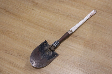 Small metal round nose shovel with white wooden handle. A twisting mechanism between metal and wood allows the item to fold.
