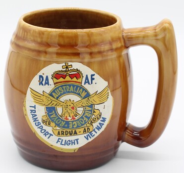 Brown stein shaped mug with handle. White painted circle on front with the RAAF emblem and the words Transport Flight Vietnam beneath that.
