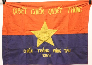 Image of rectangular flag, with red top half and dark blue bottom. A gold five-pointed star is embroidered in the middle with  messages in Vietnamese above and below..