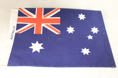 Small Australian flag with six white stars depicting the Southern Cross on blue fabric with the word Australia written horizontally on the left hand side.