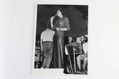 Black and white image with a long-haired woman in a long dress in the foreground. She is holding a microphone. Three males in background , two sitting and one standing back to her.