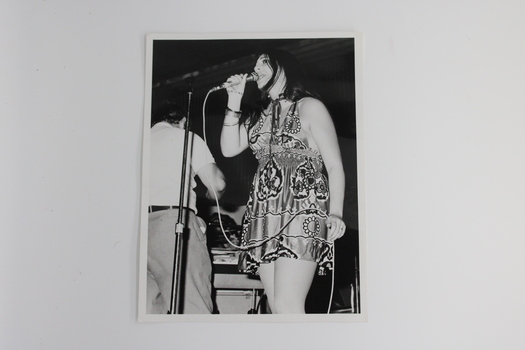 A black and white image of Linda George singing with conductor Brian may in background.