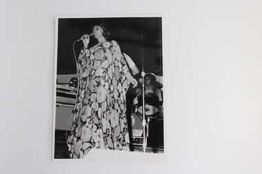 Black and white image with a female singer wearing a full-length floral kaftan in foreground.  In the background is a male seated at a drumkit.