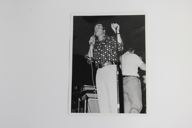 Black and white image of a male singer with handheld microphone in foreground and another male with back to him in the background. 