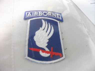 Small blue coloured US airborne shoulder patch showing a white wing and a red bayonet.