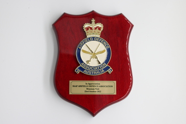 A red wooden plaque in the shape of a shield with a crown on the top of a badge with sword and rifle and an eagle.