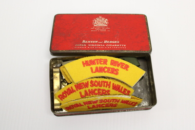 A small metal tin with a red lid with red and white writing of a cigarette brand "Benson and Hedges" Inside are a number of material epaulettes and badges.