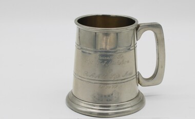 A pewter tankard with squared handle. Has a flared base and two sets of parallel lines engraved around it, plus an inscription.