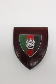 A wooden plaque with a central metal shield with green, red and black horizontal stripes and a silver badge.