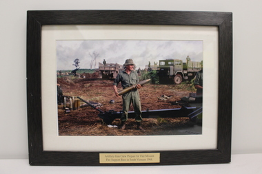 A colour image of a solider holding a mortar. In the background two heavy vehicles are shown with one soldier in each vehicle.