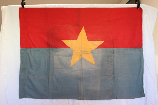 A hand made Viet Cong flag. The top half is red and the bottom half is a faded blue colour. In the centre appears a gold star. 