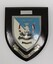 Black wooden shield with shield-shaped emblem in blue and white with a silver border. silver ribbon at bottom has a Latin motto.
