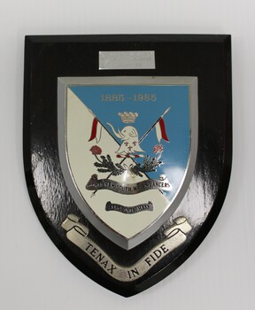 Black wooden shield with shield-shaped emblem in blue and white with a silver border. silver ribbon at bottom has a Latin motto.
